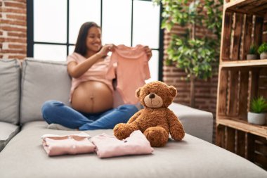 Genç Latin kadın hamile, elinde bebek kıyafetleri var.