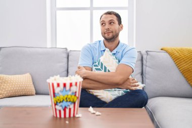 Hispanic man watching tv at home sitting on the sofa smiling looking to the side and staring away thinking. 