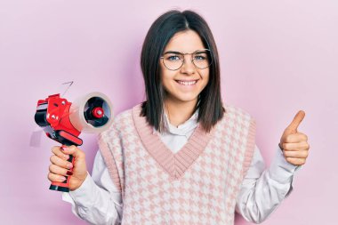 Young brunette woman holding packing tape smiling happy and positive, thumb up doing excellent and approval sign 