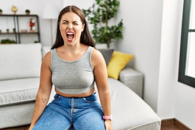 Genç esmer kadın evdeki kanepede kızgın ve kızgın bir şekilde oturmuş öfkeyle bağırıyor. öfke ve agresif konsept. 