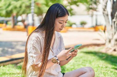 Parkta çimlerin üzerinde oturan akıllı telefon kullanan Çinli bir kadın.