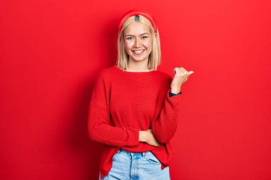 Beautiful blonde woman wearing casual red sweater smiling with happy face looking and pointing to the side with thumb up. 