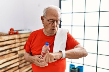 Senior man wearing sportswear and towel at the gym checking the time on wrist watch, relaxed and confident 