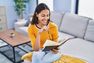 Genç Afrikalı Amerikalı kadın kitap okuyor. Evdeki kanepede oturuyor.