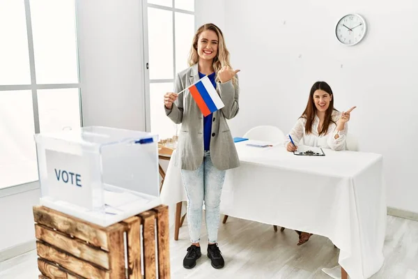 Young blonde woman at political stand holding russia flag smiling happy pointing with hand and finger to the side