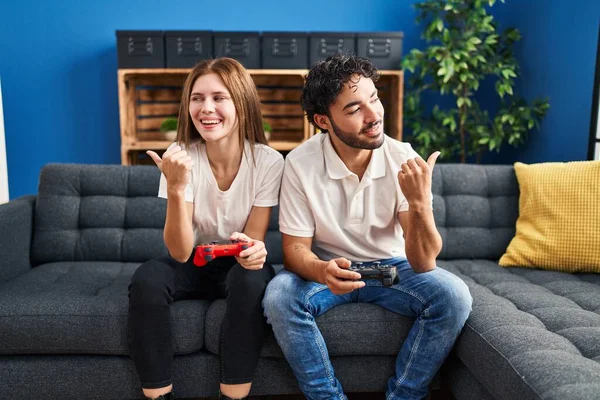 Young couple playing video game holding controller at home pointing thumb up to the side smiling happy with open mouth