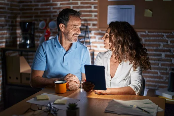 Middle age hispanic couple using touchpad sitting on the table at night looking to side, relax profile pose with natural face and confident smile.