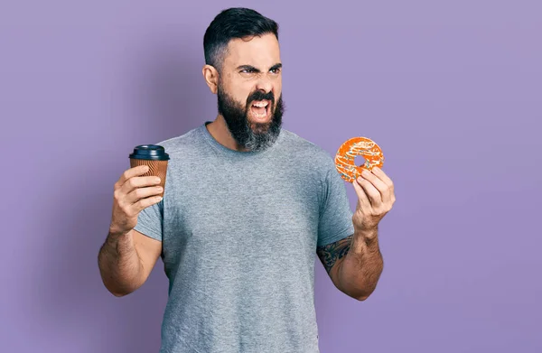 Hispanic man with beard eating doughnut and drinking coffee angry and mad screaming frustrated and furious, shouting with anger. rage and aggressive concept.