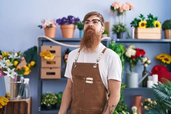 Young redhead man florist standing with serious expression at flower shop