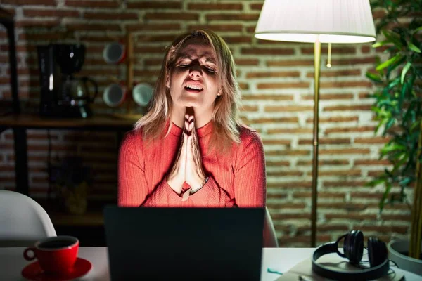 Blonde woman using laptop at night at home begging and praying with hands together with hope expression on face very emotional and worried. begging.