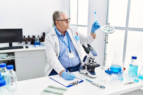 中年白发男子身穿科学家制服 手持试管 并在实验室的剪贴板上写字 — 图库照片
