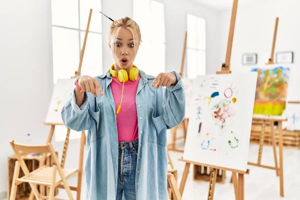Young caucasian girl at art studio pointing down with fingers showing advertisement, surprised face and open mouth