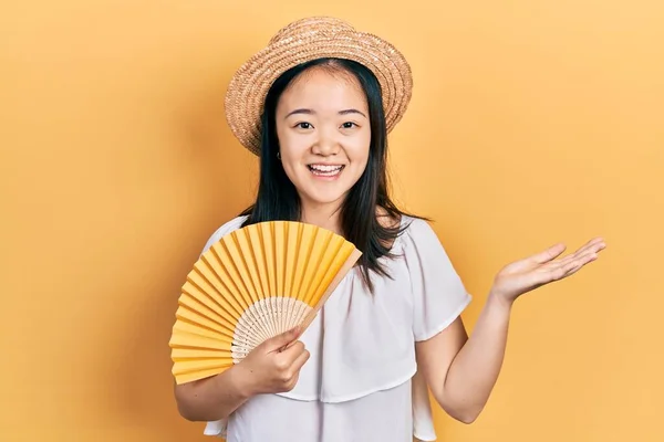 Young chinese girl waving hand fan cooling air in summer celebrating achievement with happy smile and winner expression with raised hand