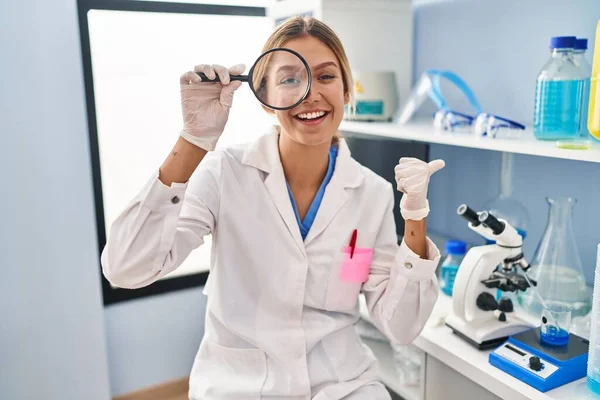 Young blonde woman working at scientist laboratory using magnifying glass pointing thumb up to the side smiling happy with open mouth