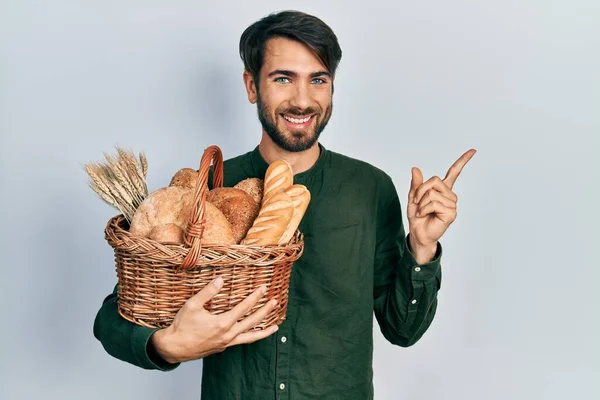 Молодий Іспаномовний Чоловік Тримає Плетений Кошик Хлібом Посміхаючись Щасливим Вказуючи — стокове фото