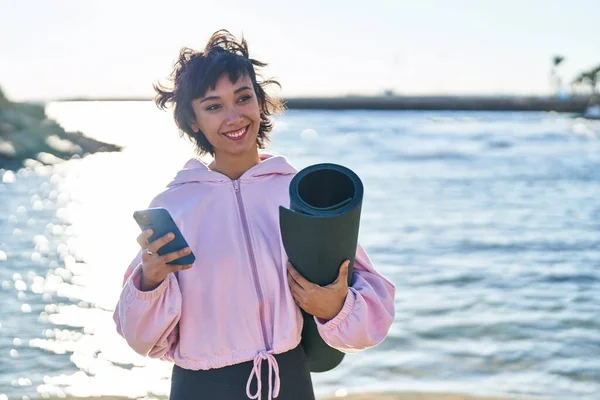 在海滨用智能手机拿着瑜伽垫的年轻女子 — 图库照片