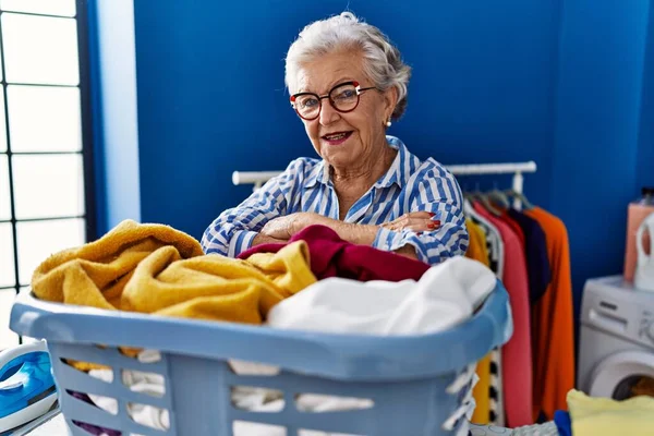 Yaşlı gri saçlı kadın gülümsüyor çamaşır odasında kıyafetlerle sepete yaslanıyor.