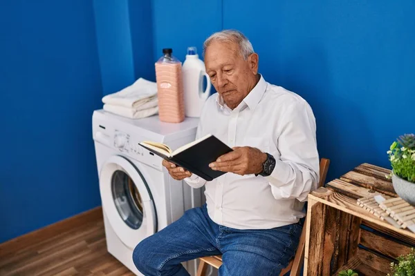 Senior man reading book waiting for washing machine at laundry room