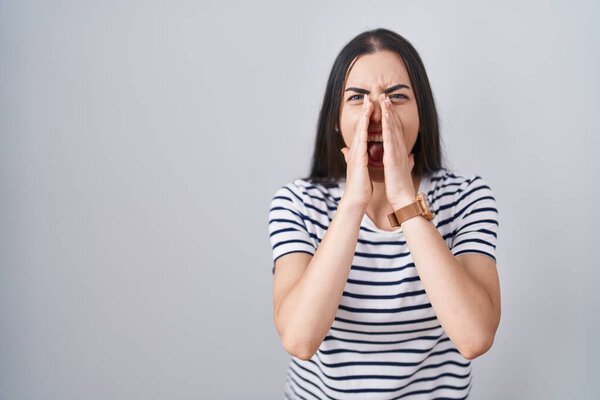 Young brunette woman wearing striped t shirt shouting angry out loud with hands over mouth 