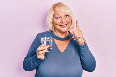 Middle age blonde woman drinking glass of water smiling with an idea or question pointing finger with happy face, number one 
