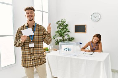 Young handsome man voting putting envelop in ballot box smiling happy and positive, thumb up doing excellent and approval sign 