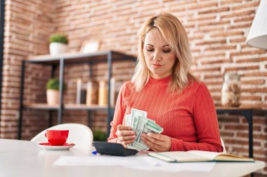 Young blonde woman sitting on table counting dollars at home
