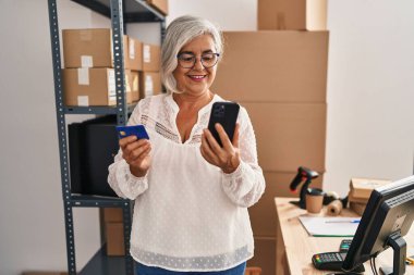 Orta yaşlı kadın ecommerce iş işçisi. Ofiste kredi kartı kullanarak akıllı telefon kullanıyor.