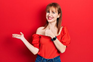Kırmızı tişörtlü kızıl saçlı genç kadın avuç içini gösteriyor ve baş parmaklarıyla iyi bir jest yapıyor, mutlu ve neşeli gülümsüyor. 