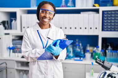African american woman wearing scientist uniform writing on clipboard laboratory