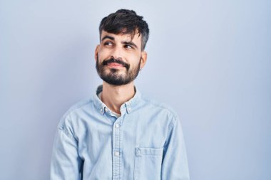 Young hispanic man with beard standing over blue background smiling looking to the side and staring away thinking. 