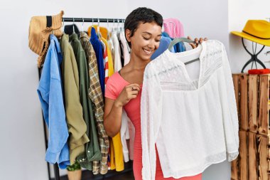 Young hispanic woman customer smiling confident choosing clothes holding shirt at clothing store
