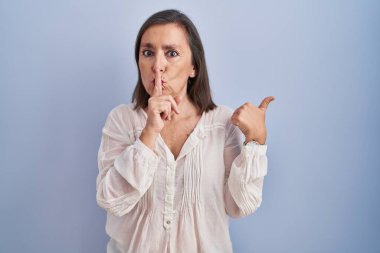 Middle age hispanic woman standing over blue background asking to be quiet with finger on lips pointing with hand to the side. silence and secret concept. 