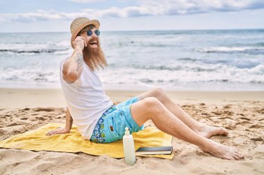 Genç kızıl saçlı adam kumsalda havlunun üzerinde oturmuş akıllı telefondan konuşuyor..