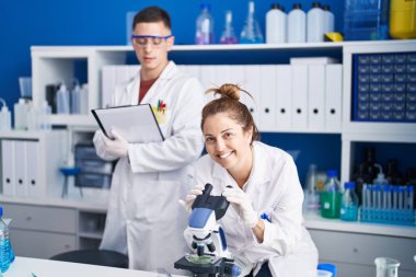 Man and woman scientists using microscope write on document at laboratory