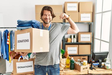 Handsome middle age man holding donations box for charity at volunteer stand strong person showing arm muscle, confident and proud of power 