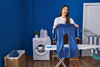 Çamaşır odasında ütü yapan esmer kadın elinde yanmış demir gömlekle kahkahalarla gülüyor.. 