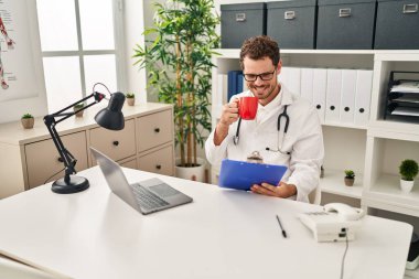 Doktor üniforması giyen, kahve içen genç İspanyol adam klinikte çalışıyor.