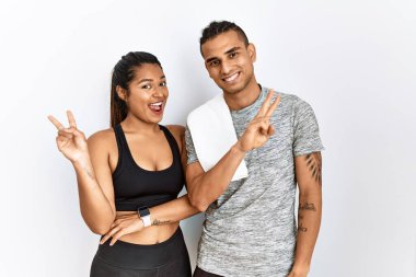 İzole edilmiş arka planda spor giyinen genç Latin çift kameraya bakıp gülümsüyor ve zafer işareti yapıyor. İki numara.. 