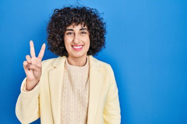 Young brunette woman with curly hair standing over blue background showing and pointing up with fingers number two while smiling confident and happy. 