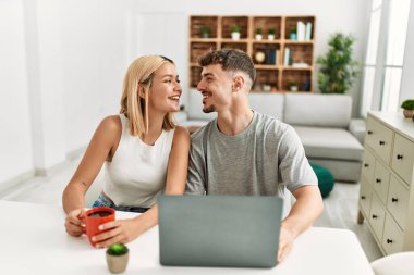 Genç beyaz çift dizüstü bilgisayar kullanıyor ve kahve içiyor. Evdeki masada oturuyorlar..