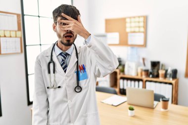 Hispanic man with beard wearing doctor uniform and stethoscope at the office peeking in shock covering face and eyes with hand, looking through fingers with embarrassed expression. 