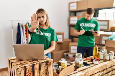 Young woman and man wearing volunteer t shirt at donations stand with open hand doing stop sign with serious and confident expression, defense gesture 