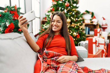 Genç Latin kadın akıllı telefonun yanında selfie çekiyor. Yılbaşı ağacının yanında oturuyor.