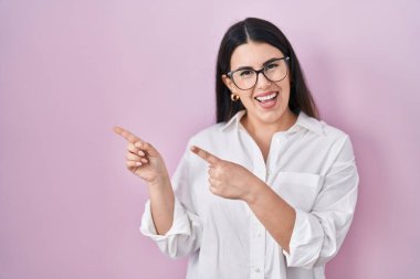 Young brunette woman standing over pink background smiling and looking at the camera pointing with two hands and fingers to the side. 