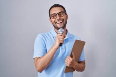 Hispanic man holding reporter microphone and clipboard smiling and laughing hard out loud because funny crazy joke. 
