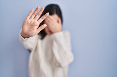 Genç esmer kadın mavi arka planda duruyor ellerle gözleri örtüyor ve üzgün ve korku ifadeleriyle dur hareketi yapıyor. utangaç ve olumsuz kavram. 