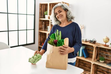 Orta yaşlı, gri saçlı, elinde poşetlerle evde bekleyen bir kadın..