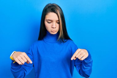Young brunette girl wearing turtleneck sweater pointing down looking sad and upset, indicating direction with fingers, unhappy and depressed. 