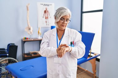 Middle age woman with grey hair working at pain recovery clinic checking the time on wrist watch, relaxed and confident 