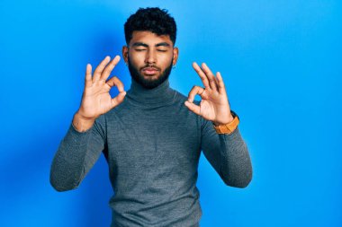 Arab man with beard wearing turtleneck sweater relaxed and smiling with eyes closed doing meditation gesture with fingers. yoga concept. 